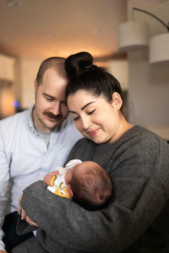 Mom and dad sitting close together looking down at their newborn son snuggled in his mom's arms for Gig Harbor in-home newborn session