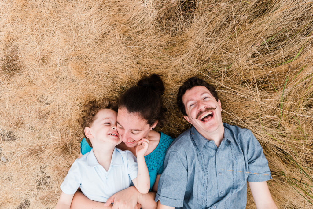 Family laying in tall grass looking up at the camera with genuine smiles for family photos